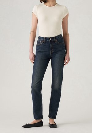 Vrouw staand, gekleed in een nauwsluitend wit T-shirt met korte mouwen, donkere rechtvallende jeans met een hoge taille en zwarte balletschoenen, tegen een neutrale achtergrond.