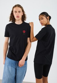 Černé bavlněné trička s vyšívanou červenou růží na hrudi, nošené s světle modrými džíny a černými šortkami. Minimalistický design.