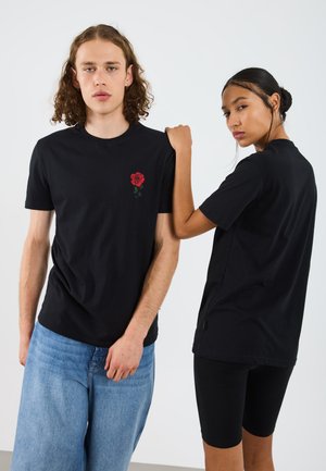 T-shirt di cotone neri con ricamo di una rosa rossa sul petto, indossati con jeans azzurri chiari e pantaloni corti neri. Design minimalista.