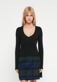 Ξανθιά γυναίκα που φοράει μαύρο μακρυμάνικο τοπ με V λαιμόκοψη και μπλε-πράσινη καρό φούστα, στέκεται μπροστά σε ένα απλό λευκό φόντο.