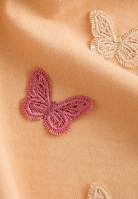 Pfirsichfarbener Stoff mit pink und cremefarbenen gestickten Schmetterlingen. Aufwändige Stichdetails auf den Flügeln, weiche Textur und glatte Oberfläche.