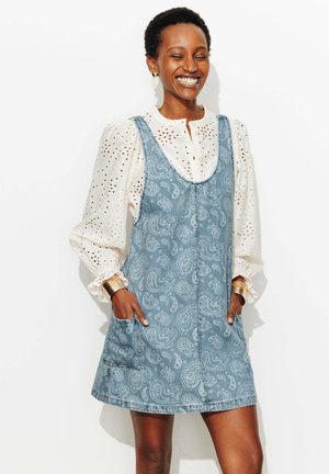 Femme souriante aux cheveux courts portant un chemisier en broderie anglaise blanche et une robe salopette en denim à motif cachemire bleu, mains dans les poches, debout devant un fond blanc.