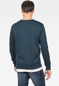 Man i mörk teal långärmad sweatshirt över vit skjorta och blå jeans, stående vänd bort mot en vit bakgrund.