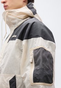 Αντιανεμικό μπουφάν με μπεζ και μαύρο χρωματικό συνδυασμό, με φερμουάρ σε τσέπη στο μανίκι και ψηλό γιακά. Η υφή φαίνεται ελαφριά.