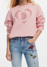 Vaaleanpunainen collegepaita, jossa on rei'itetty sydäntyyli, yhdistettynä vaaleansinisiin farkkuihin, joissa on punaisia kukkakirjontaa vasemmalla jalalla.