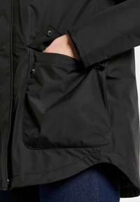 Svart vattentät jacka med en stor ytterficka med tryckknappsstängning. Slät textur, skräddarsydd design och rundad nederkant.