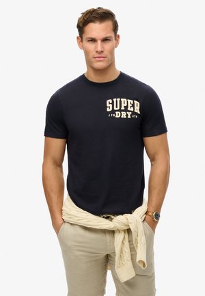 T-shirt in cotone blu navy con logo "SUPER DRY" in crema, scollatura rotonda, maniche corte; pantaloni corti khaki; maglione in crema annodato in vita.