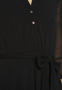 Schwarzes Kleid mit V-Ausschnitt und zwei silbernen Knöpfen, featuring einen Stoffgürtel, der in einer Schleife an der Taille gebunden ist, und transparenten langen Ärmeln.