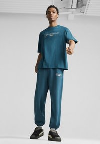 Mėlynas sportinis komplektas, kuriame yra laisvas marškinėliai su užrašu "Sportswear" ir kelnes su elastiniais rankogaliais, demonstruojamas su juodais ir pilkais sportbačiais.