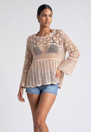 Vrouw draagt een beige gehaakte top met lange mouwen over een zwarte bikinitop en blauwe denimshorts, poseert met één hand op de heup tegen een lichte achtergrond.