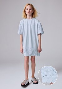 Světlomodré šaty s vyšívaným vroubkovaným lemem, krátkými rukávy a jemným texturovaným vzorem. Kombinované se šedými sandály s dekorativními kovovými prvky.