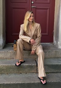 Beżowy garnitur dopasowany z ostrymi klapami, spodnie z szerokimi nogawkami; model siedzi na kamiennych schodach, ma na sobie czarne klapki, obok minimalistyczna torba.