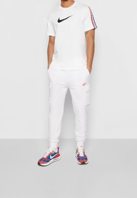 Vit Nike-t-shirt med svart logga och röd, blåa ränder på ärmarna, ihop med vita cargobyxor med fickor och färgglada sneakers.