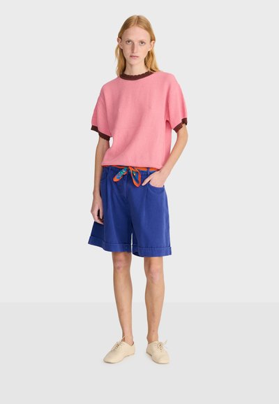 Jeune femme debout portant un haut rose à manches courtes, un short bleu jusqu'aux genoux avec une ceinture écharpe colorée, et des chaussures beiges.