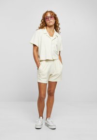 Hellbeige, kurz geschnittenes Hemd mit Knopfverschluss und passende Shorts aus weichem Material. Getragen mit weißen Sneakers und rosa getönten Sonnenbrillen.