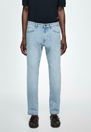 Mann trägt hellblaue Jeans mit geradem Bein, kombiniert mit schwarzen Lederschuhen und einem schwarzen kurzärmligen Oberteil, steht vor einem schlichten Hintergrund.