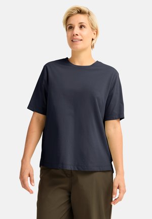 Femme aux cheveux blonds courts portant un t-shirt bleu foncé ample et un pantalon marron, debout devant un fond clair uni.
