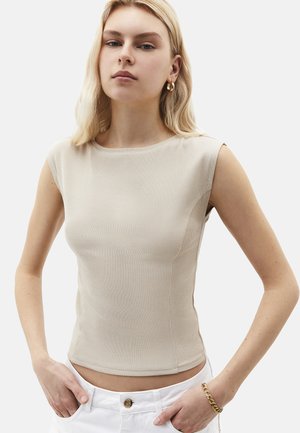 Jeune femme portant un haut sans manches beige à côtes, un pantalon blanc avec détail de boutons, des boucles d'oreilles créoles dorées et un bracelet doré, debout les mains dans les poches.