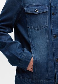 Τζάκετ από τζιν σε βαθύ μπλε χρώμα με ξεθωριασμένο εφέ. Περιλαμβάνει μπροστινές τσέπες, κλείσιμο με κουμπιά και ορατές λεπτομέρειες ραφής.
