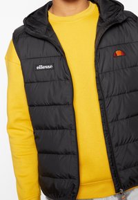 Svart vadderad väst med huva, med dragkedjestängning och en logotyp-patch, lagrad över en gul sweatshirt med rund hals.