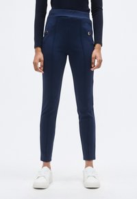 Σκούρο μπλε παντελόνι ψηλόμεσο από ελαστικό ύφασμα, με δύο μπροστινές τσέπες διακοσμημένες με μεταλλικά κουμπιά και λεπτή γραμμή εφαρμογής.