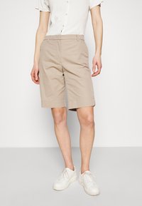 Beige maßgeschneiderte Shorts aus einem Baumwollmischgewebe mit flacher Vorderseite, Seitentaschen und leicht umgeschlagenen Säumen, kombiniert mit weißen Sneakern.