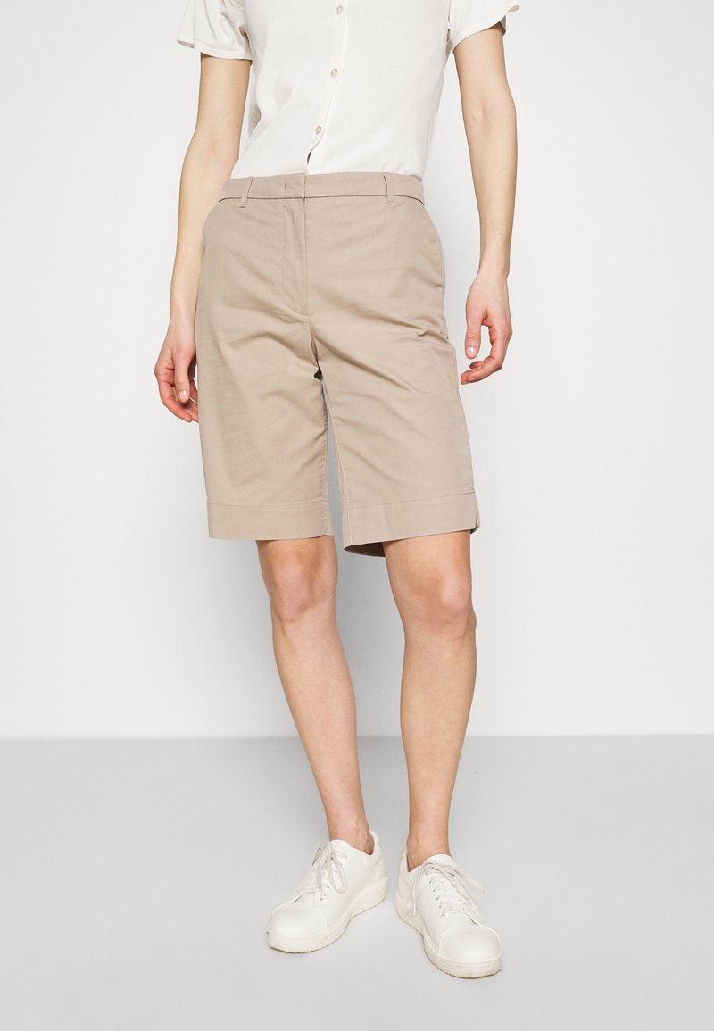 Beige maßgeschneiderte Shorts aus einem Baumwollmischgewebe mit flacher Vorderseite, Seitentaschen und leicht umgeschlagenen Säumen, kombiniert mit weißen Sneakern.