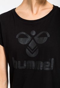 Černé bavlněné tričko s velkým, reflexním šedým logem "hummel" a stylizovaným emblémem nahoře. Kolo výstřih a krátké rukávy.