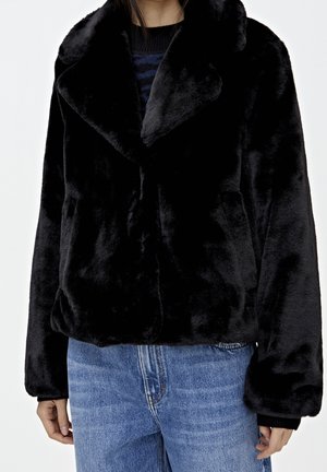 Woman wearing a black faux fur jacket with large collar over a dark sweater and blue denim jeans, standing against a white background.