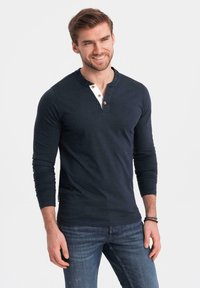 Maglietta henley a maniche lunghe blu navy con colletto bianco e tre bottoni. Indossata con jeans blu, caratterizzati da un tessuto testurizzato e un design aderente.