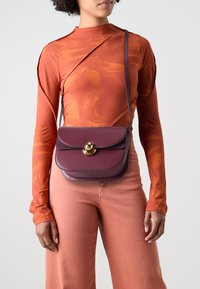 Borsa a tracolla in pelle bordeaux con chiusura dorata, forma tondeggiante e superficie liscia. Indossata a tracolla sopra una maglietta arancione a maniche lunghe.