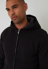 Strikket sort zip-up hoodie med et struktureret mønster, udstyret med en foret hætte og sølvhardware. Foran lynlås lukning og ribbede manchetter.