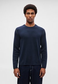 Tumši zila adīta džemperis ar garām piedurknēm, apaļu izgriezumu kaklā un smalku kontrastējošu šuvju detaļu pie plecu šuvēm. Gluda tekstūra.