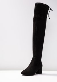 Bota negra de ante por encima de la rodilla con tacón de bloque, que presenta una textura suave y un detalle de cintas en la parte posterior para un ajuste ajustable.