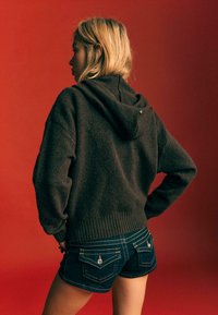 Brauner Kapuzenpullover aus strukturiertem Stoff, mit geripptem Saum und lockerer Passform, kombiniert mit dunkelblauen Denim-Shorts, die dekorative Nähte und Akzente aufweisen.