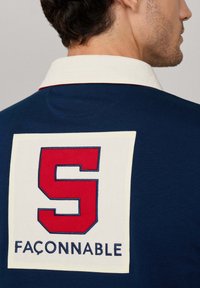 Navy-blaues Polohemd mit einem off-weißen Kragen. Auf der Rückseite befindet sich ein großes rotes 'S' und das Wort 'FAÇONNABLE' in Blau. Weicher Stoff.