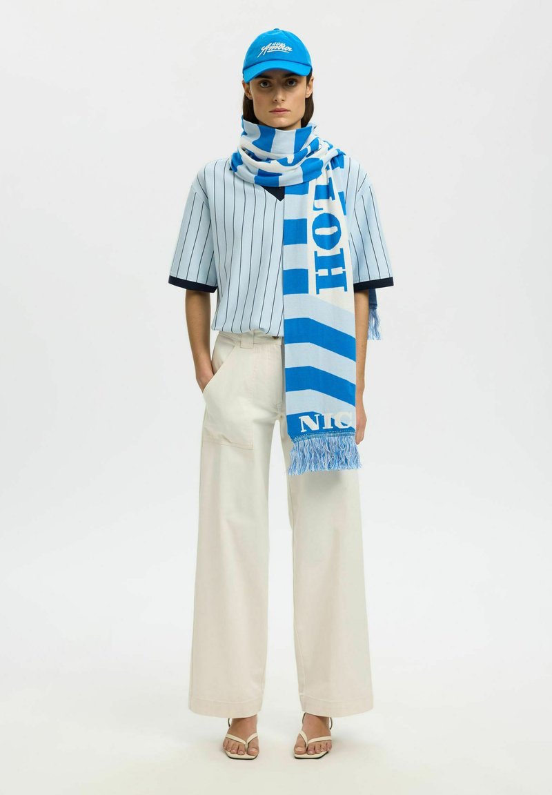 Femme portant un pantalon blanc à jambes larges, un t-shirt de baseball rayé, une écharpe bleue et blanche, une casquette bleue et des sandales beiges, debout les mains dans les poches.
