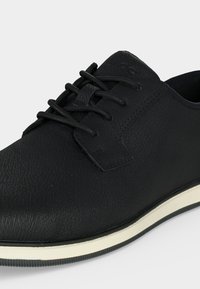 Chaussure noire en cuir texturé à lacets avec semelle en caoutchouc blanche et noire, gros plan montrant les détails avant et côté.