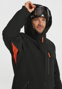 Zwarte winterjas met een capuchon, voorzien van oranje accenten en ritssluitingen, textuurstof en meerdere ritszakken.