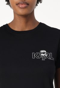 Svart bomull t-skjorte med et hvitt tegneserieillustrasjon av en karakter og teksten "KARL" i fet skrift på venstre bryst.