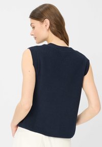 Donna con capelli castani che indossa un gilet a maglia senza maniche blu navy e pantaloni bianchi, vista di spalle e leggermente di lato.