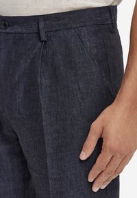 Mörkblå linnebyxor i texturerat tyg, med framvikta veck, knappstängning och smal passform, visade på en hand.