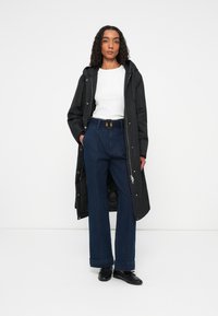 Zwarte waterdichte parka met capuchon, voorzien van een ritssluiting en drukknoopsluiting aan de voorkant. Witte getailleerde blouse, hoogwaardige blauwe wijde broek en zwarte schoenen.