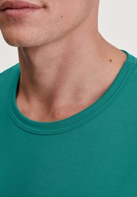 Teal crewneck t-shirt gemaakt van zachte, gestructureerde stof. Close-up van de halslijn en bovenste borst toont gladde huid en kleine onvolkomenheden.