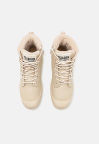 Paire de bottes Palladium beige avec des lacets épais, des fermetures éclair latérales et une doublure en polaire moelleuse visible à l'ouverture supérieure.