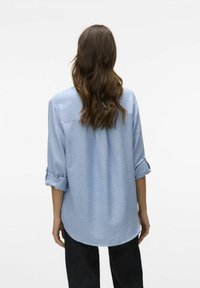 Svetlo modra, dolg rokav, majica s zavihanimi manšetami, okroglo robom in mehkim materialom. Zadnji pogled prikazuje sproščen kroj in minimalne detajle.
