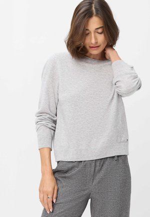 Vrouw met kort bruin haar die een lichtgrijze trui en grijze geruite broek draagt, kijkt naar beneden terwijl ze met één hand haar nek raakt.