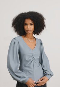 Blusa grigia chiara con scollo a V, centro arricciato e maniche a sbuffo. Realizzata in tessuto morbido, con una texture liscia e un design aderente.