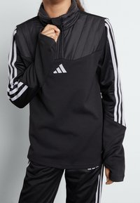 Czarna sportowa bluza z wysokim kołnierzem, pikowanym przednim panelem oraz długimi rękawami z białymi paskami i logo. Miękka, teksturowana tkanina.