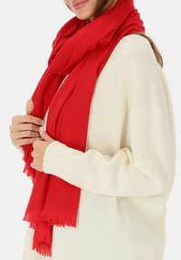 Czerwona szalik z miękkiego materiału, z frędzlami, owinięty wokół szyi na kremowym swetrze z dzianiny.